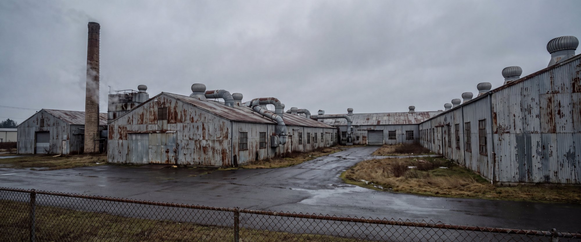 A disused industrial plant of rusting metal buildings and ventilation ducts under a grey sky
